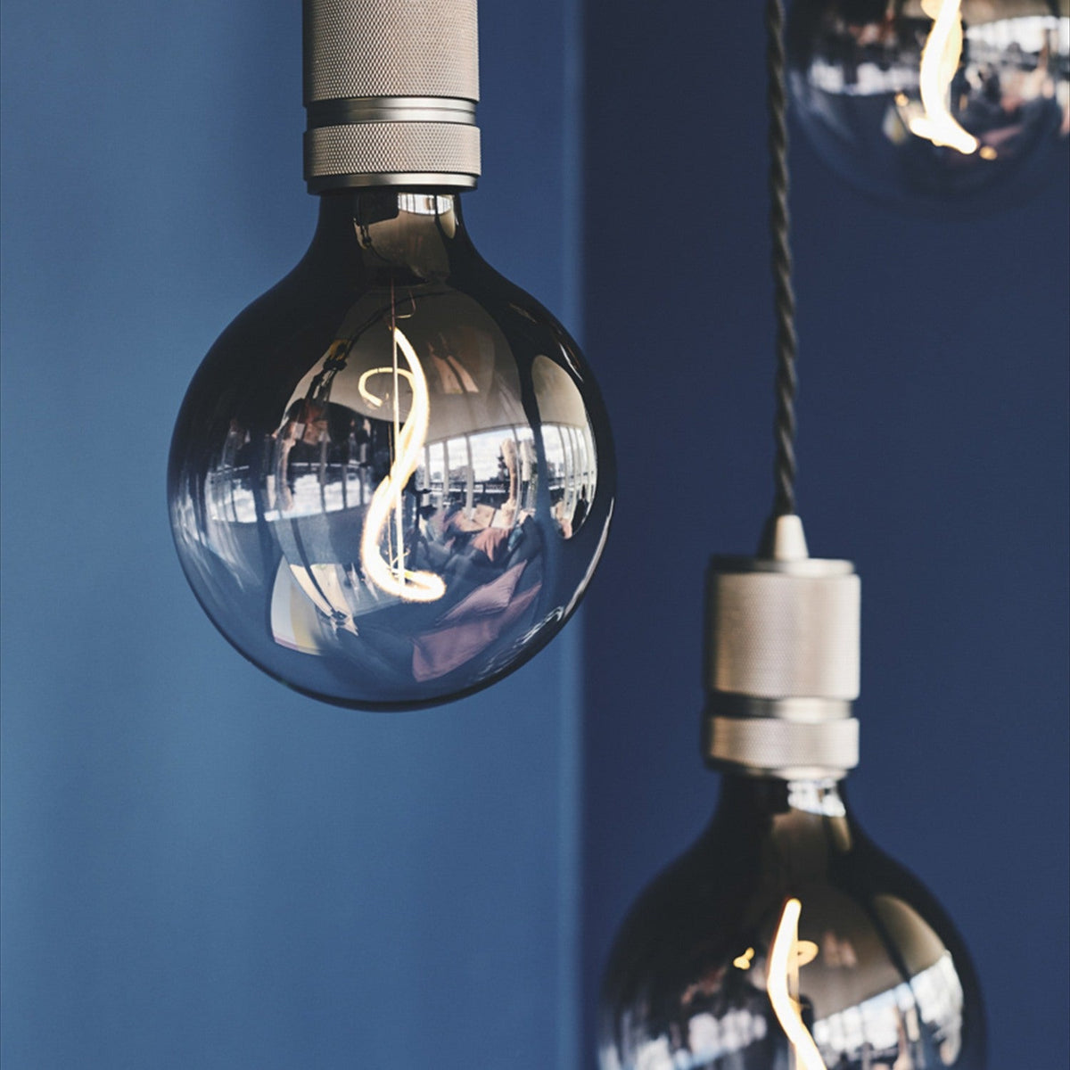 Halo Cable Cord Set E27 pendant lampholders in gunmetal with knurled detailing and twisted black flex, shown lit with globe bulbs against a dark blue wall.