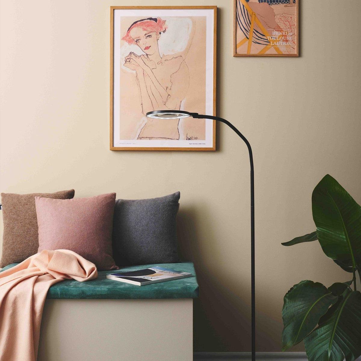 Magni LED X3 Dioptri Floor Lamp in black with a curved slim stem, round magnifying lens head and flat disc base, shown beside a bench with cushions in a living room.