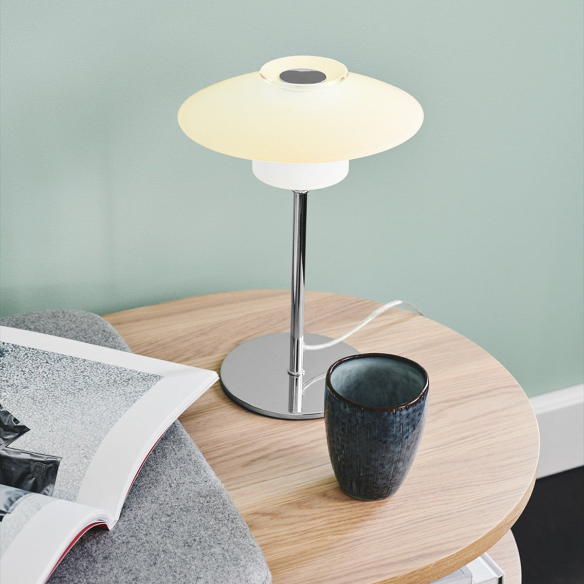 Scandinavia Table Lamp 20cm in opal glass and chrome, shown lit on a wooden side table beside a ceramic cup and magazine against a sage green wall.