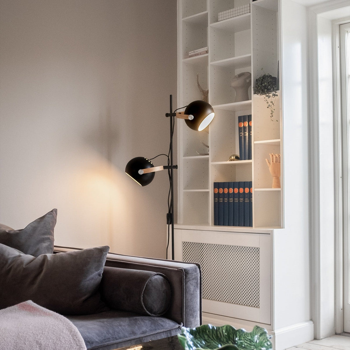 DC 2 Light Floor Lamp in black and oak, with a slim stem and two adjustable black spheres on oak arms, shown beside a bookcase in a living room.