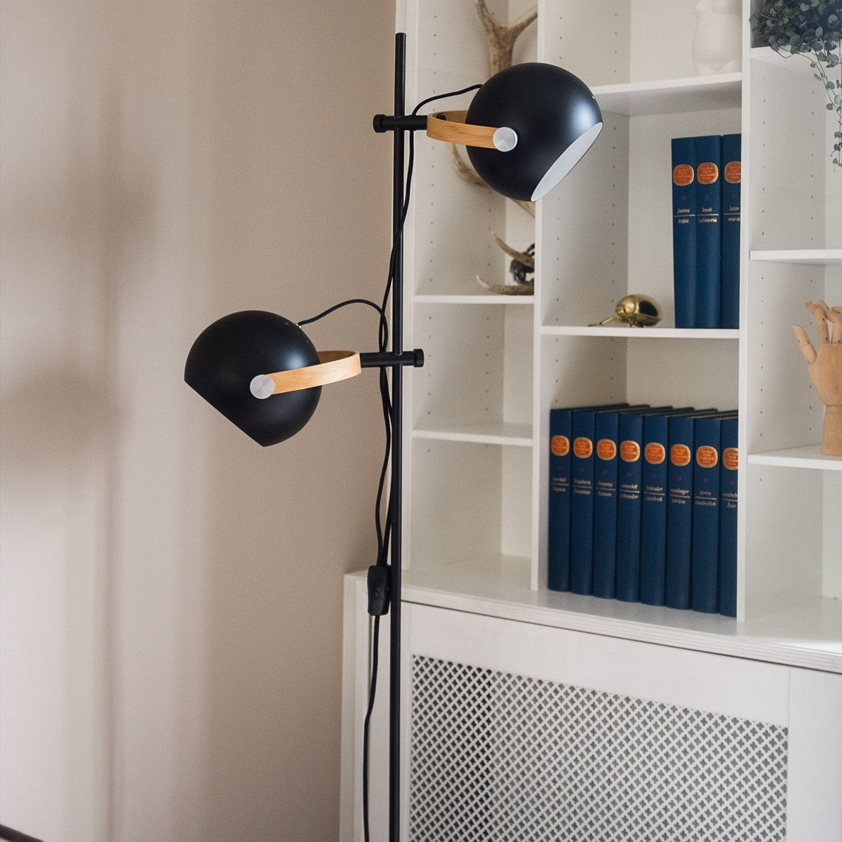 DC 2 Light Floor Lamp in black and oak, with two adjustable black spherical heads on oak arms and a slim black stem, shown beside a white bookshelf.