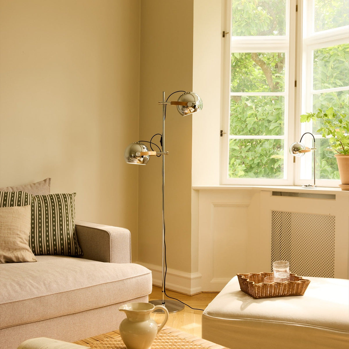 DC 2 Light Floor Lamp in chrome with two adjustable sphere shades on oak arms, shown in a living room beside a window.
