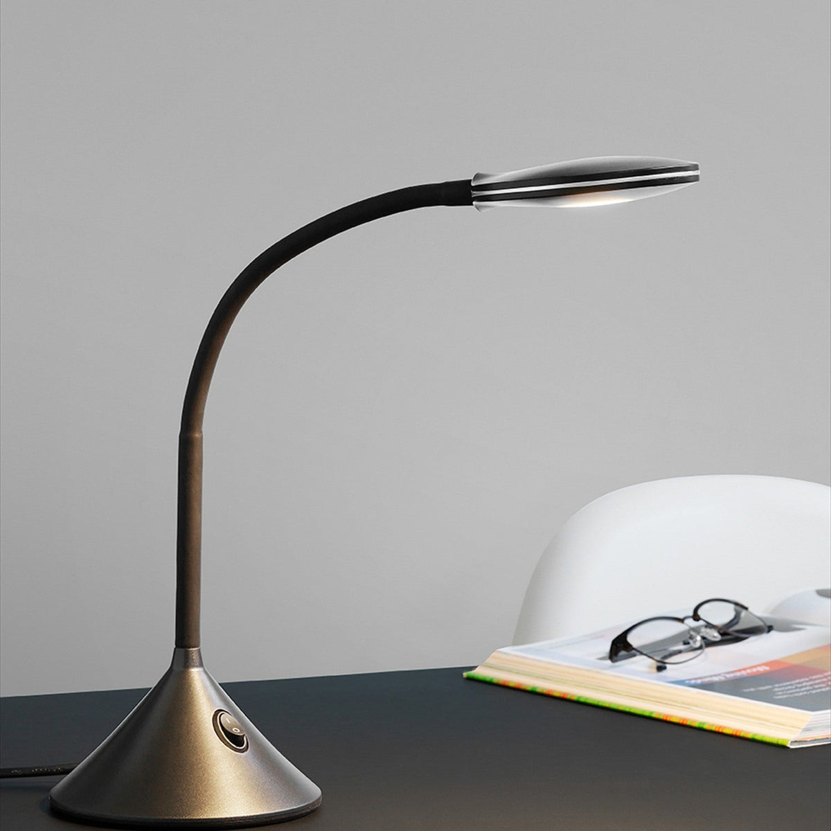 Fix LED Table Lamp 4W in black, with a curved flexible arm, oval head, and gunmetal cone base with a round switch, shown on a desk beside glasses and a book.