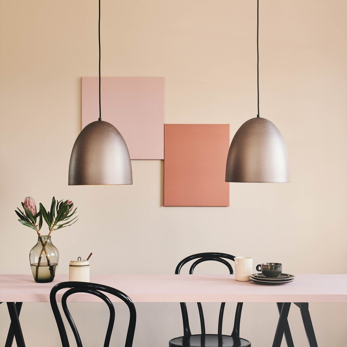 Two Classic Oxid Brushed Pendant Lights in bronze, dome-shaped with a wide open rim, hung above a pink dining table with black chairs.