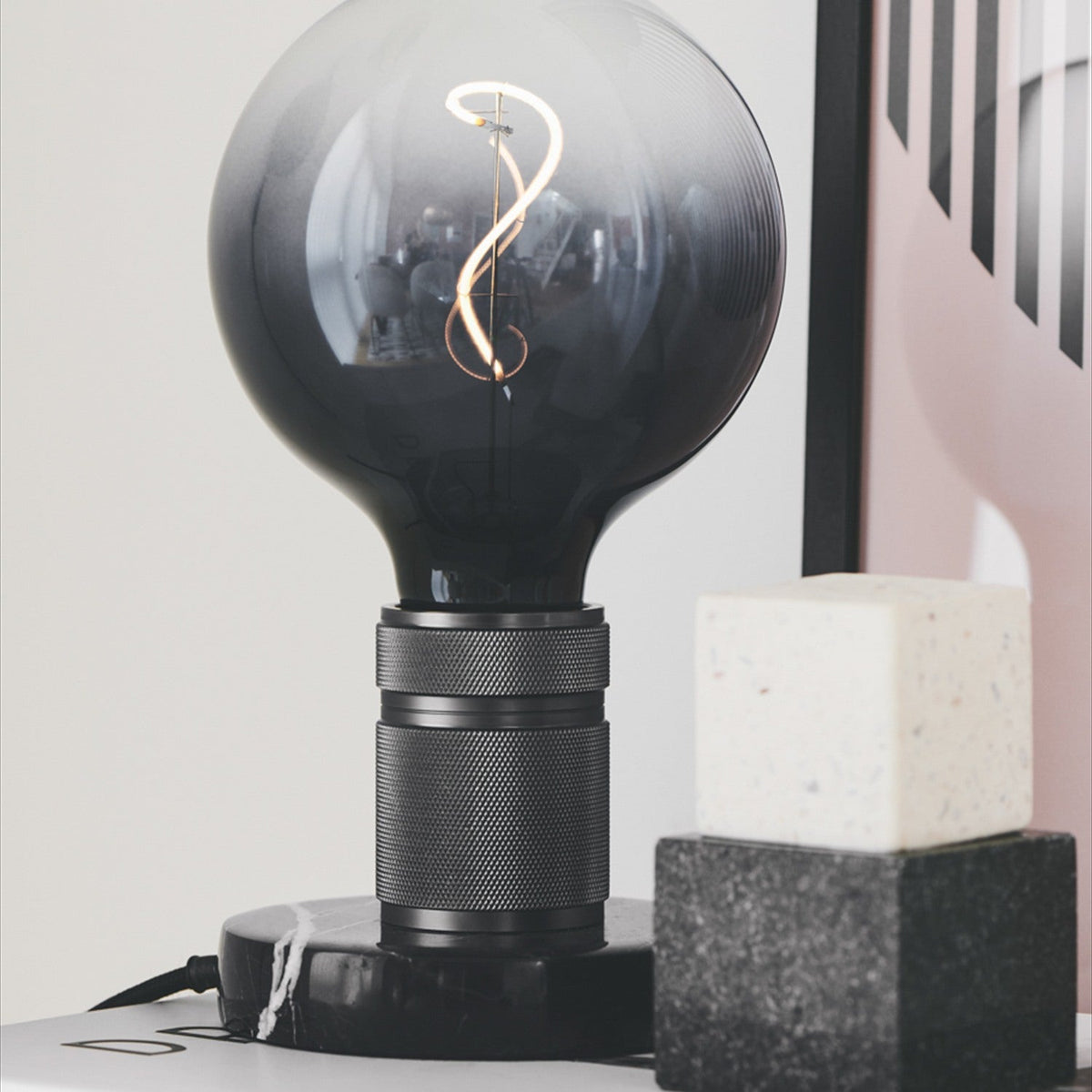 Halo Table Lamp in gunmetal and marble, knurled cylindrical base with black veined marble disc, fitted with a large smoked globe bulb, styled near decor blocks.