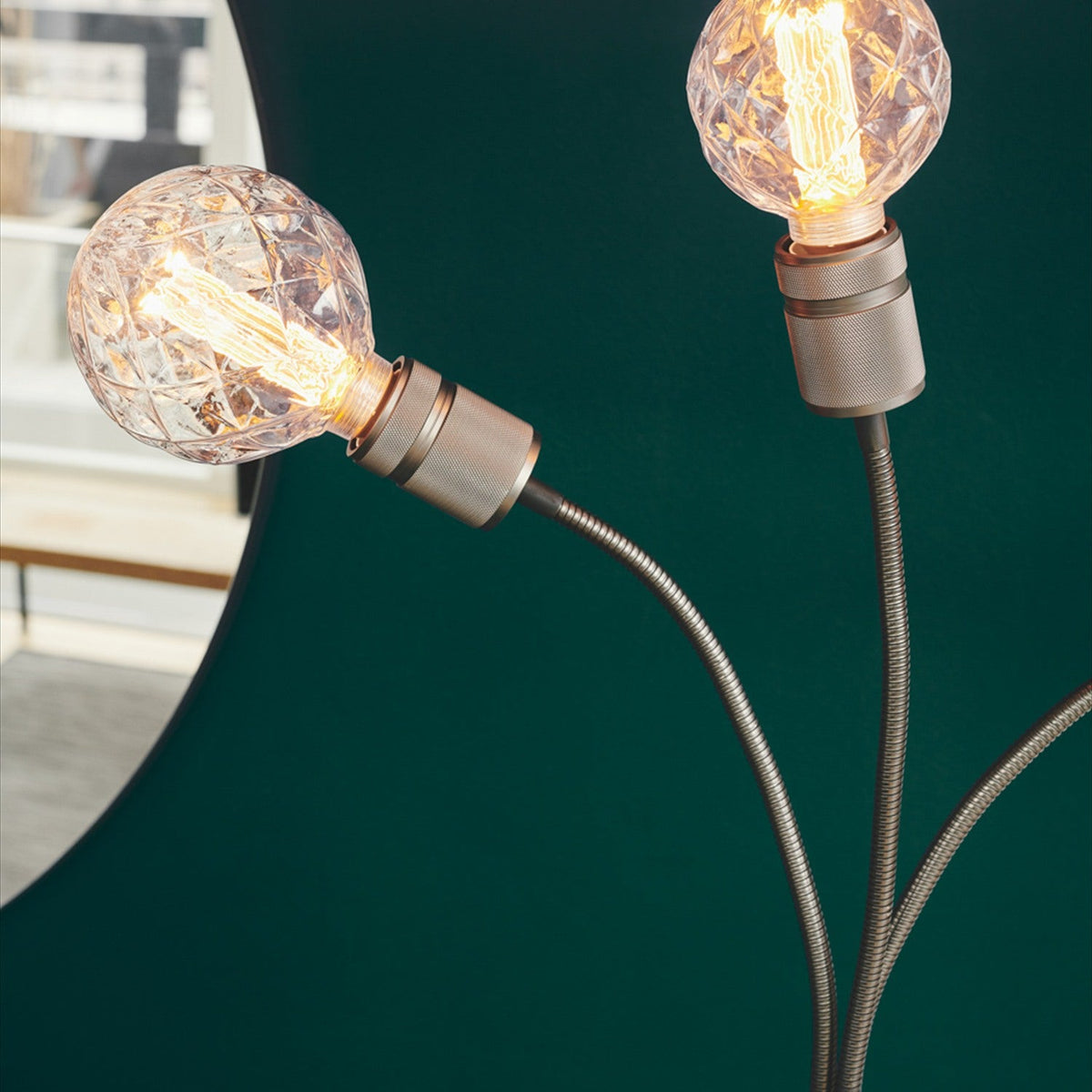Halo 3 Light Floor Lamp in gunmetal, close-up of two curved flexible arms with knurled holders and textured glass globes lit with visible filaments against a dark green backdrop.