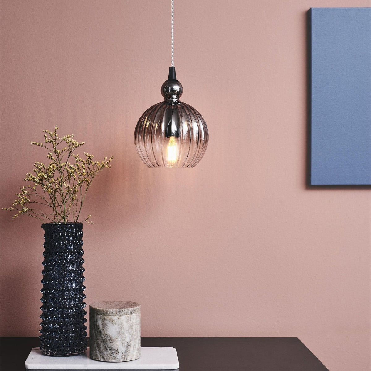 Ball Pendant Light 15cm in smoke ribbed glass with black cone fitting, hanging above a dark table with vase and marble pot against a pink wall.