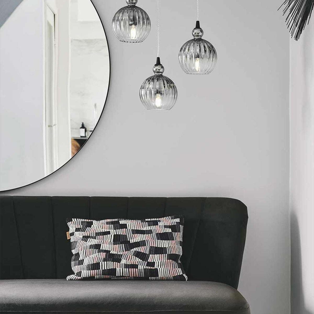 Ball Pendant Light 15cm in smoke, three ribbed smoked-glass globes with black cone fittings hung at staggered heights above a dark upholstered sofa near a round mirror.
