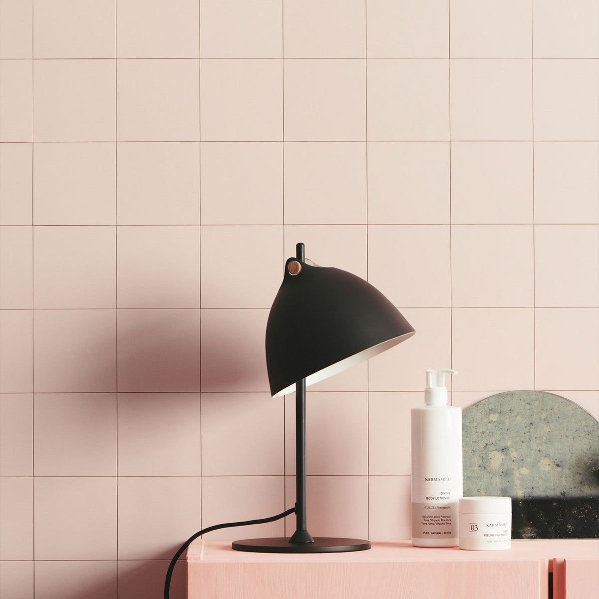 Aarhus Table Lamp 18cm in black and wood, angled dome shade with cream interior on a slim black post and round base, shown on a pink cabinet against pink tiled wall.