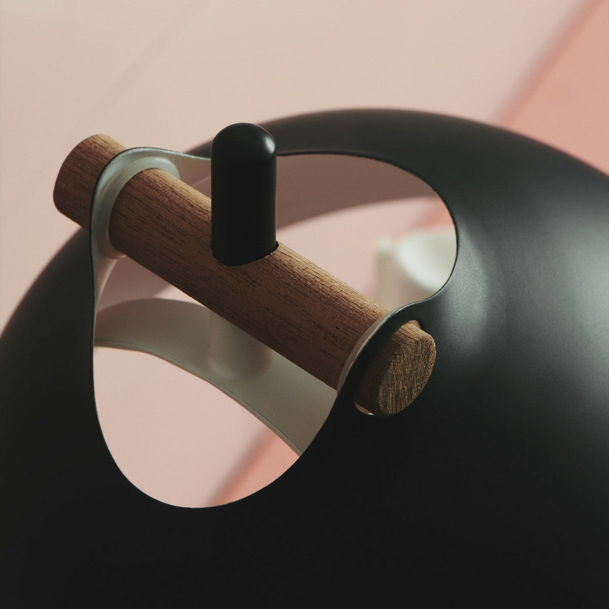 Close-up of the Aarhus Table Lamp 18cm in black and wood, showing the black dome shade's cream-lined interior, wooden handle, and black post against a pink background.