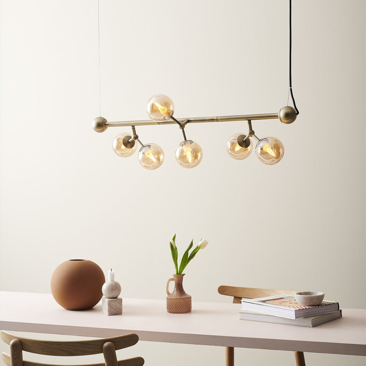 Atom Mini Horizontal 6 Light Chandelier in antique brass with amber glass spheres, hung above a dining table styled with vases and decor.