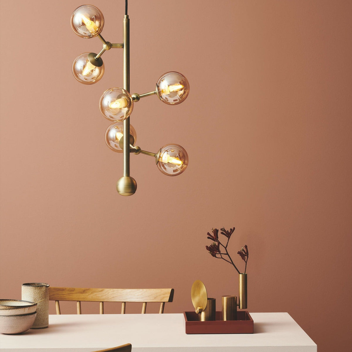 Atom Vertical 6 Light Chandelier in antique brass with six amber glass globes branching up a vertical rod, shown lit above a dining table against a terracotta wall.