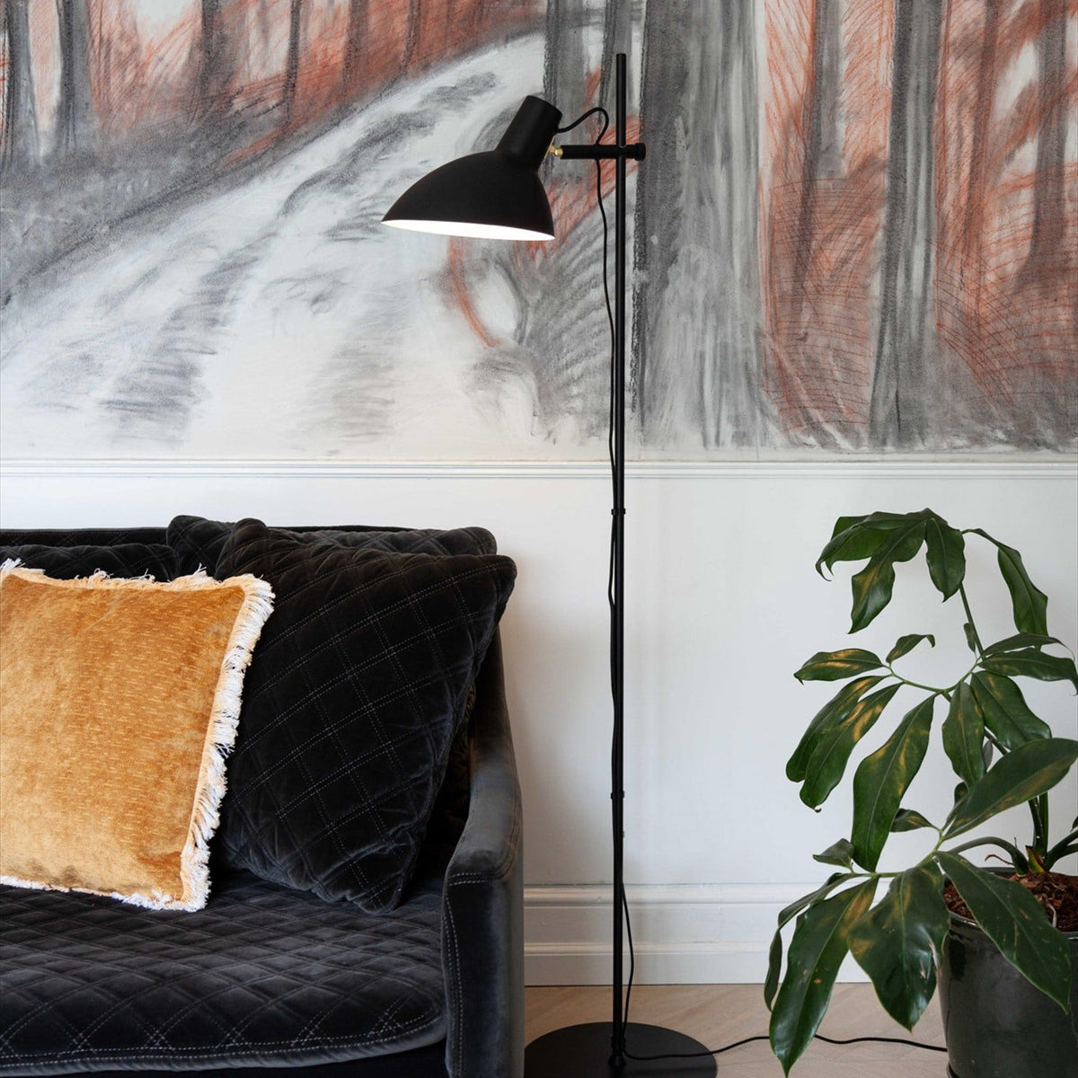 Metropole black floor lamp with slim post, round disc base, and adjustable dome shade, shown beside a velvet sofa with mustard cushion and potted plant.