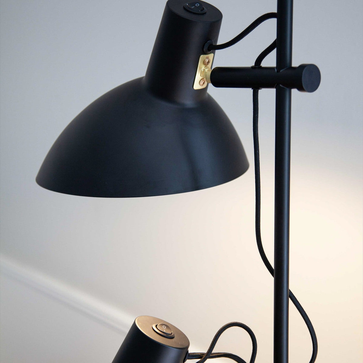 Close-up of the Metropole 2 Light Floor Lamp in black, showing a shallow dome shade with brass mounting bracket, on/off toggle switch, and fabric cable against the matte black pole.
