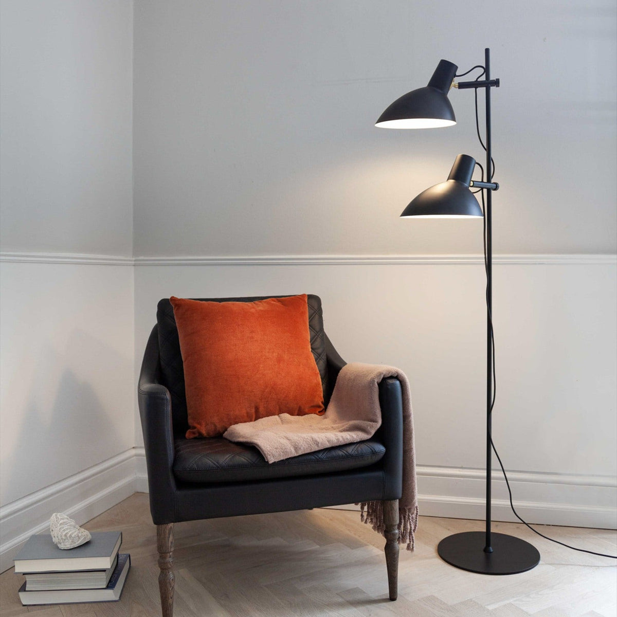 Metropole 2 Light Floor Lamp in black, with slim post, round disc base, and two adjustable dome shades at different heights, shown lit beside a leather armchair.