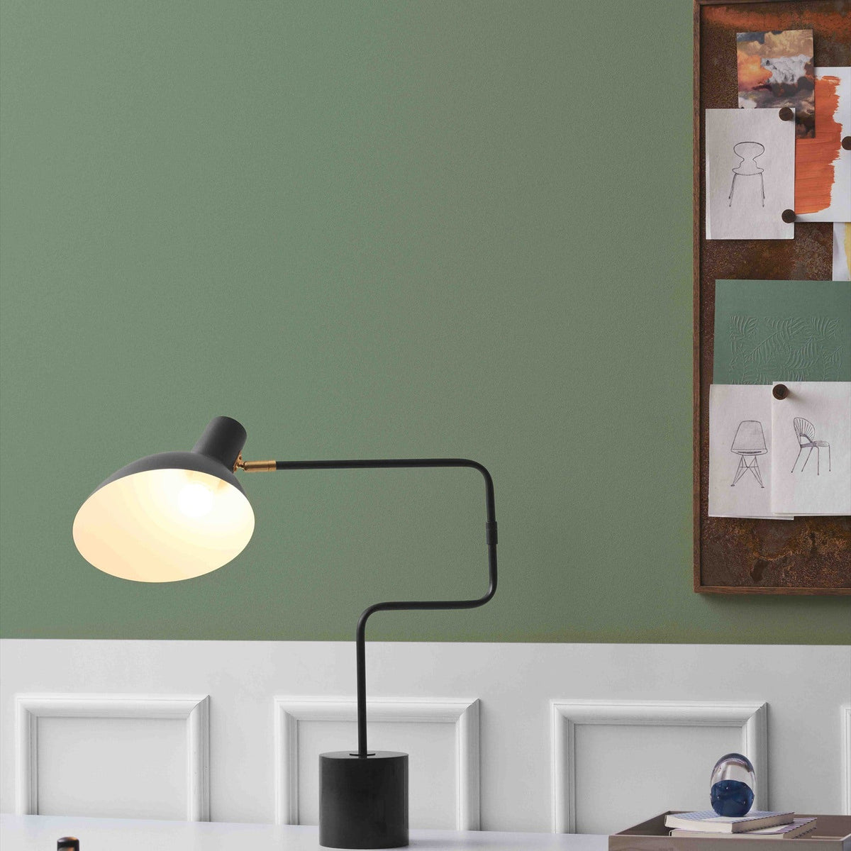 Metropole Deluxe Swing Table Lamp in anthracite with a wide dome shade, angled black arm and black marble base, shown lit on a desk against a green wall.