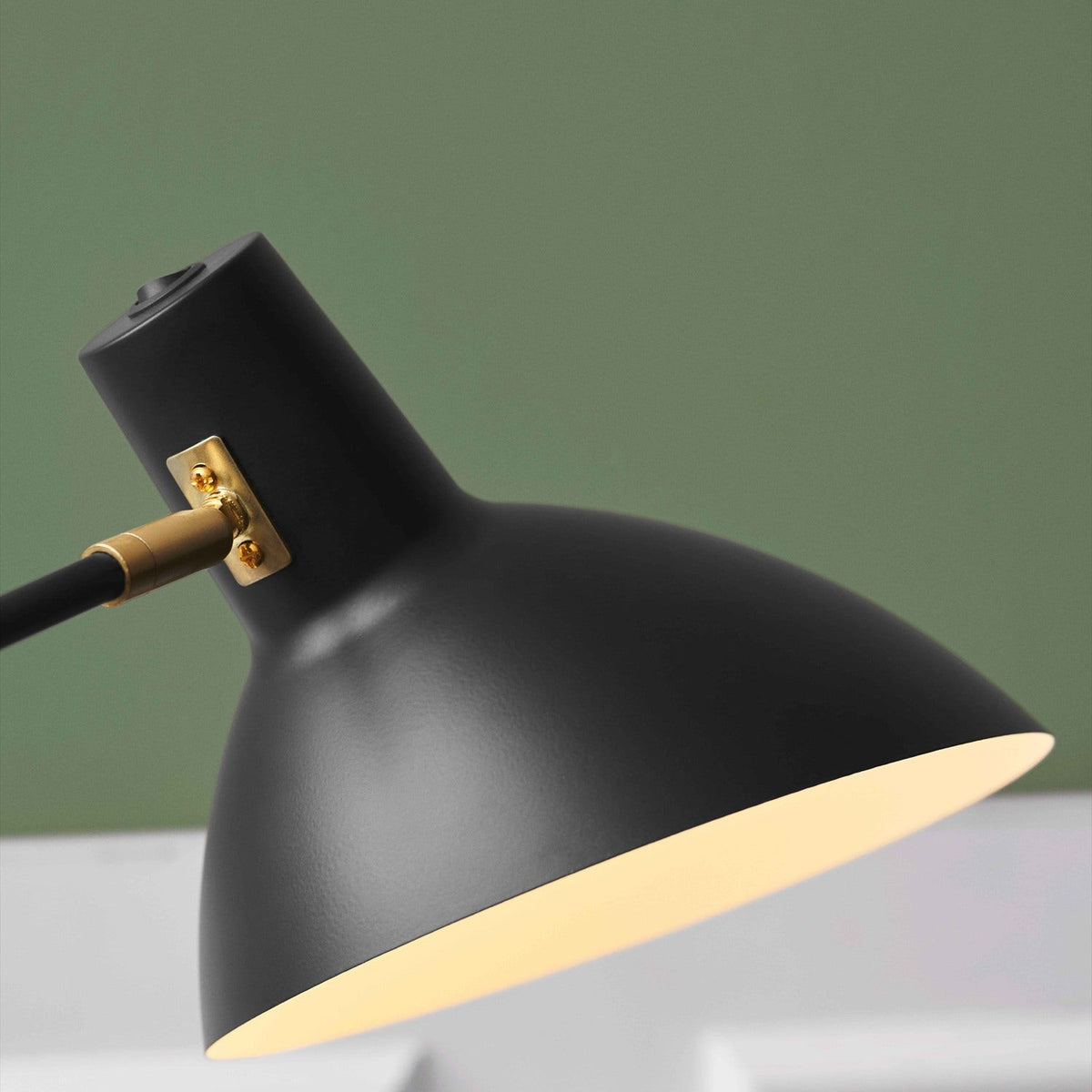 Close-up of the Metropole Deluxe Swing Table Lamp's wide, shallow anthracite dome shade with pale interior lining, brass knuckle joint, and black arm, against a green backdrop.