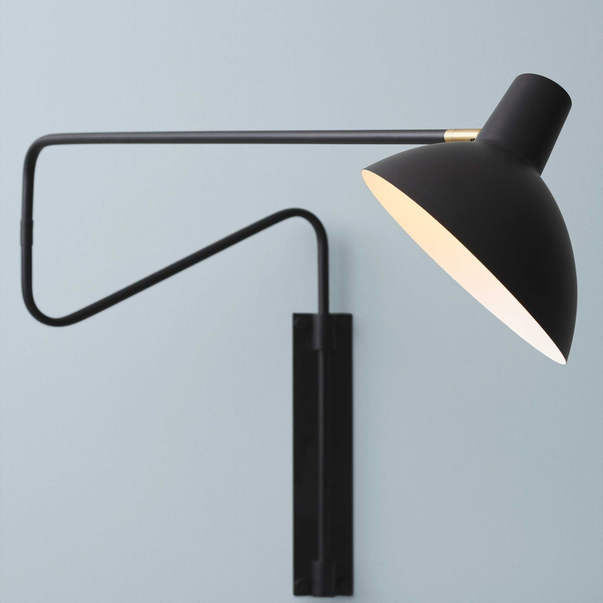 Metropole Deluxe Swing Wall Light in black, featuring a wide dome shade with pale interior, brass knuckle collar, and a folding jointed arm on a wall-mounted plate against a light grey background.
