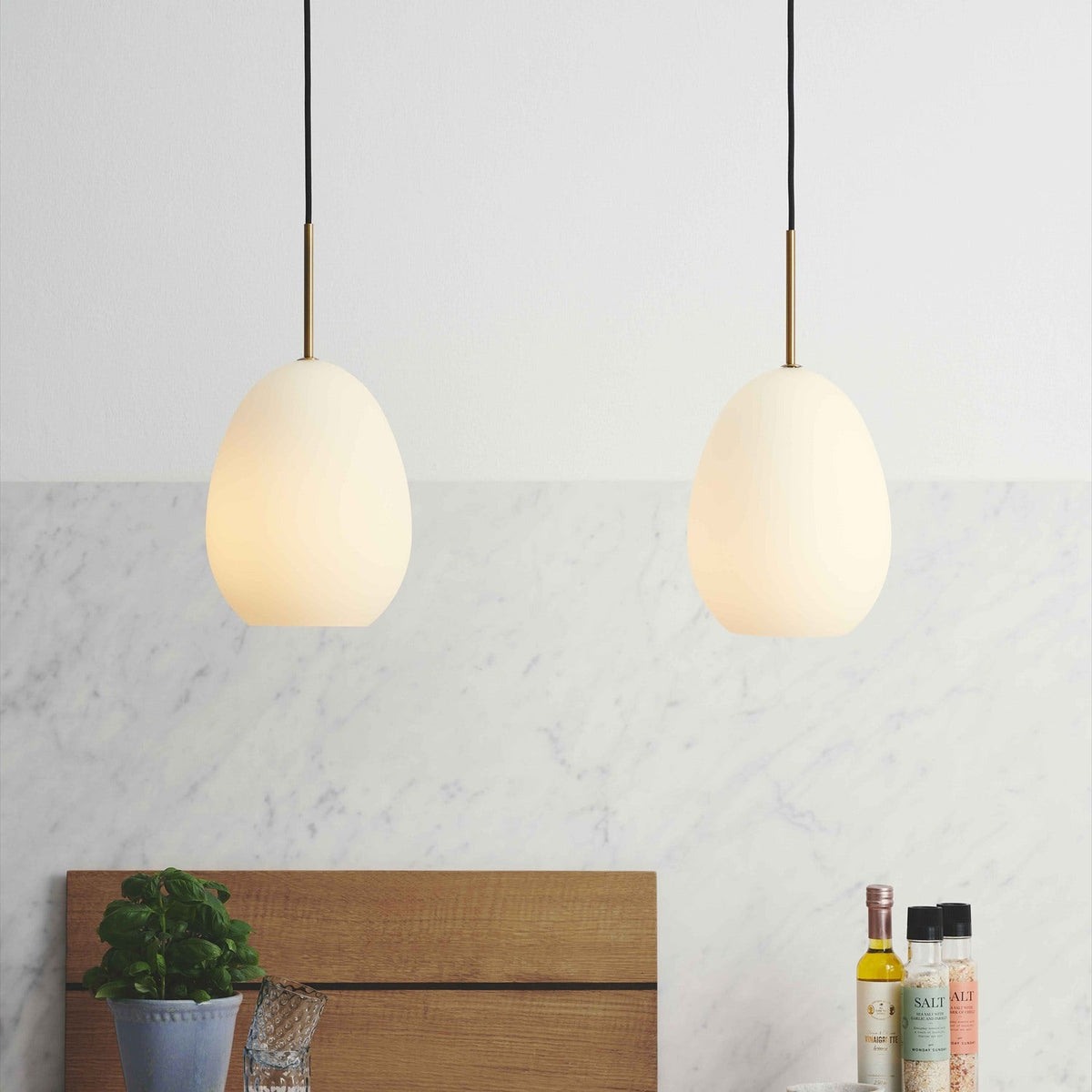 Bodø Pendant Light 20cm in brass and white, two egg-shaped opal glass shades on brass necks with black flex, lit above a wooden shelf near a marble wall.