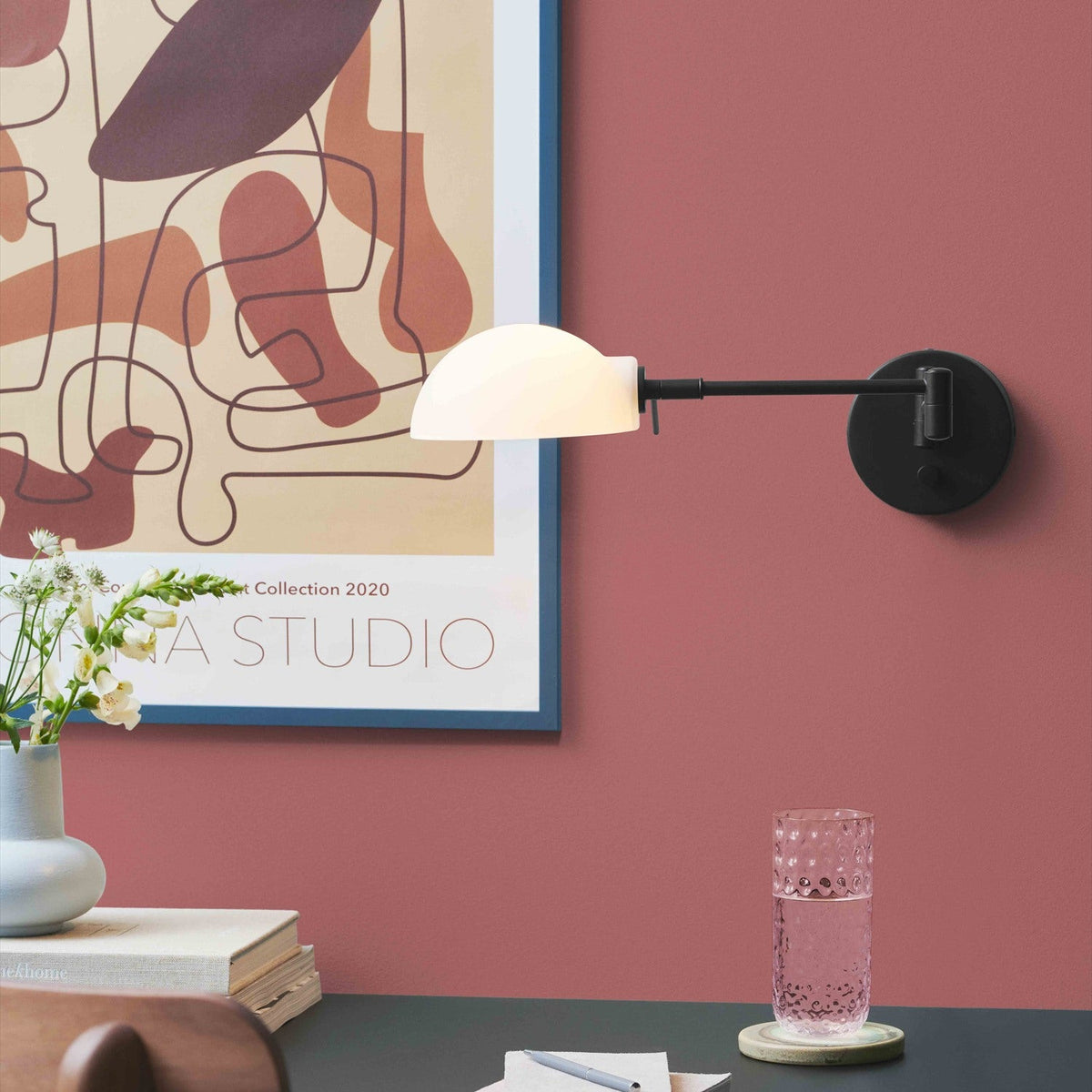 Copenhagen Wall Light in black, with a round wall plate, jointed extending arm and white half-dome shade, shown mounted above a desk against a red wall.