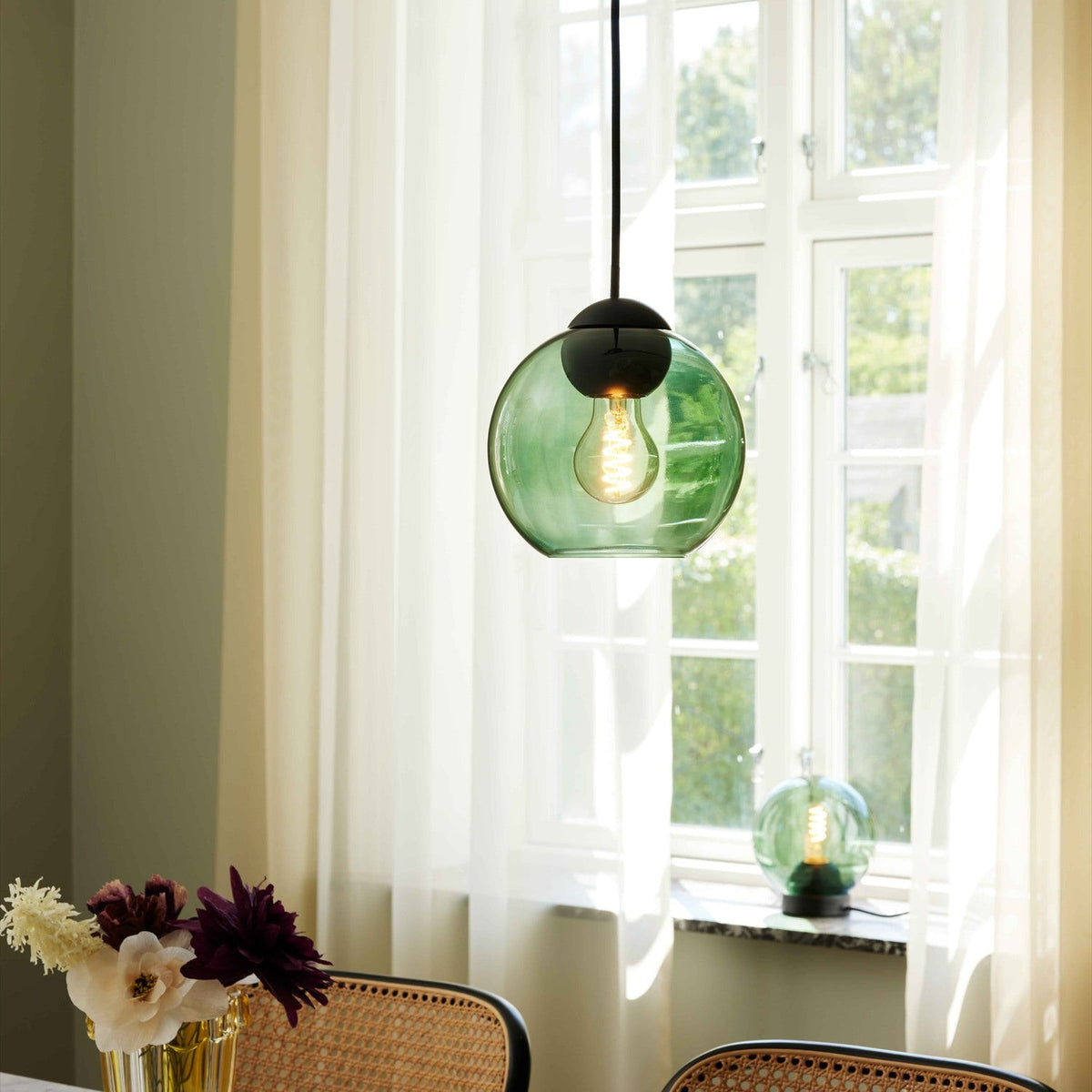 Bubbles Pendant Light 18cm in green glass with black cap fitting and visible E27 bulb, shown hanging in a dining room near a sheer-curtained window.