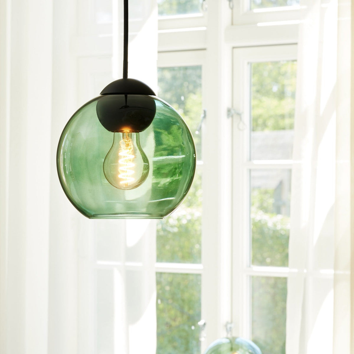 Bubbles Pendant Light 18cm in green, a round green glass globe with black cap and flex, showing a glowing E27 bulb, hung near a bright window.