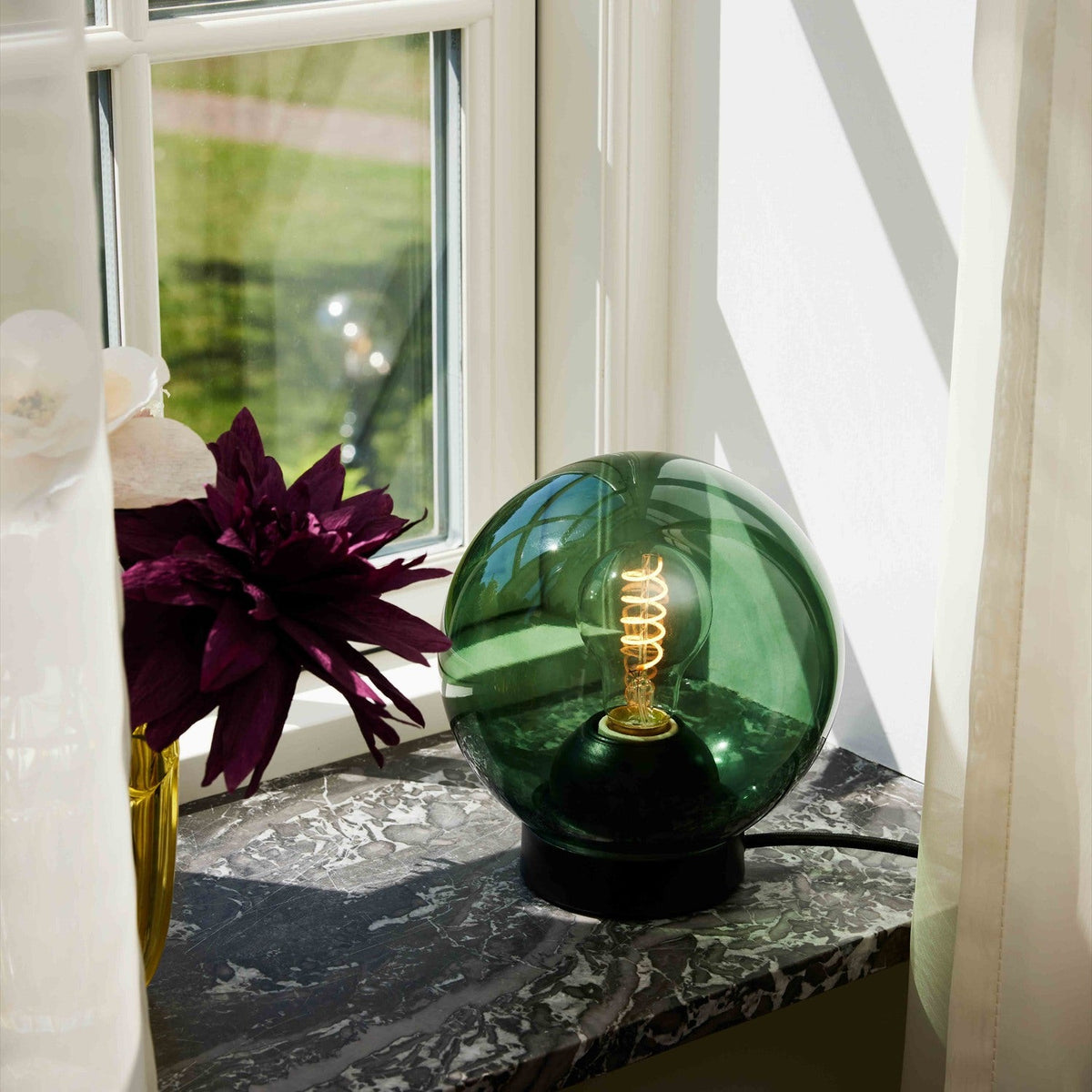 Bubbles Table Lamp 18cm in green, a round clear-tinted glass globe on a black cylindrical base with visible spiral bulb, shown on a marble windowsill beside flowers.