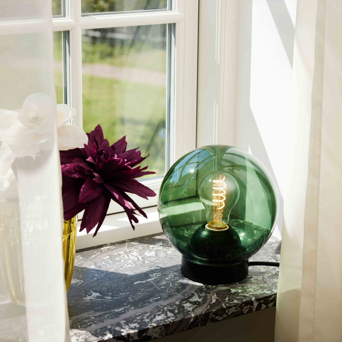 Bubbles Table Lamp 18cm - Green: a round green glass globe on a black cylindrical base with visible spiral bulb, shown on a marble windowsill.