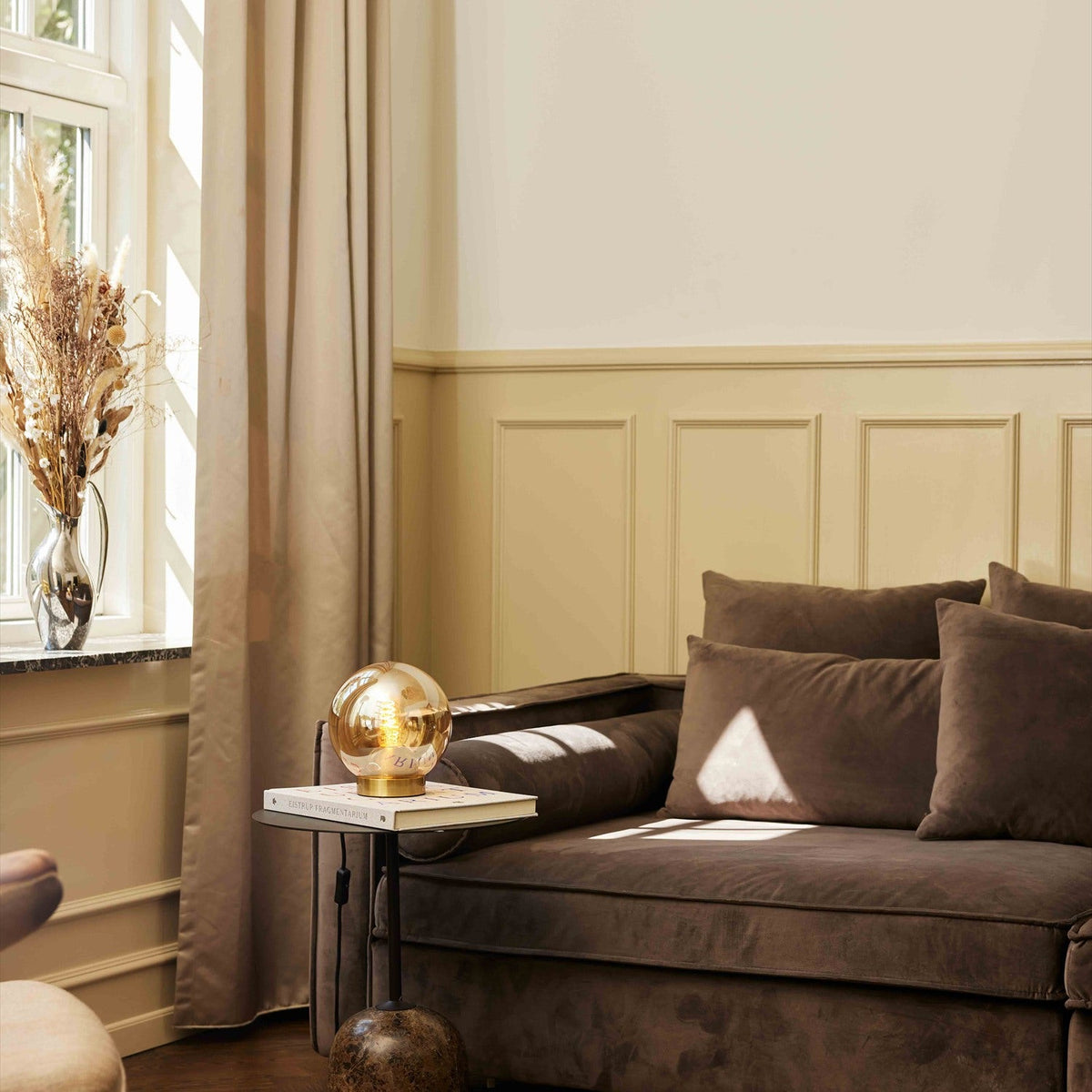 Bubbles Table Lamp 18cm in amber glass with a brass base, resting on a side table beside a brown velvet sofa in a living room setting.