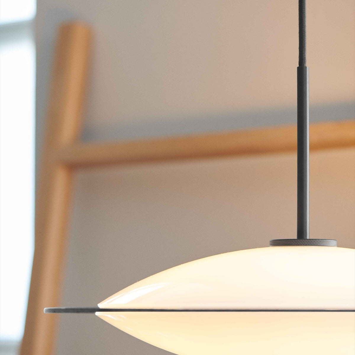Close-up of the Orbit Glass 3 Light Pendant Light 40cm in Opal & Black, showing the opal glass dome set in a thin black disc with black cord, against a softly blurred wooden ladder in the background.
