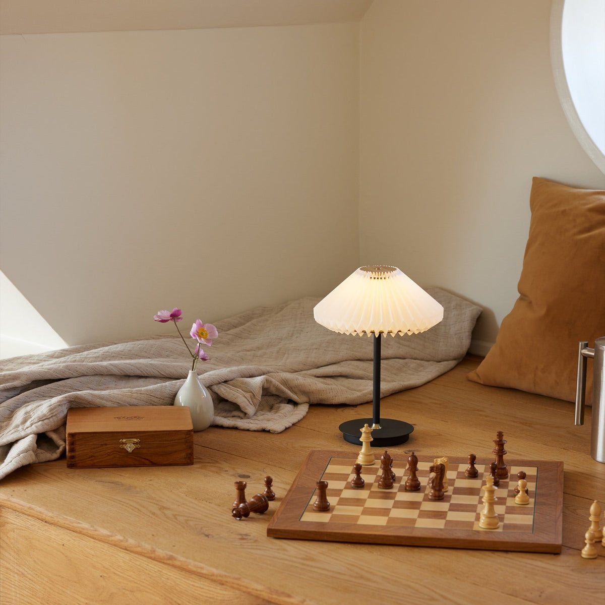 Paris LED Go Clean Rechargeable Table Lamp 22cm in white and black, with a pleated fan-style shade on a slim black stem, styled in a cosy living room beside a chessboard and linen throw.