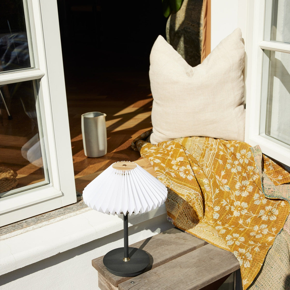 Paris LED Go Clean Rechargeable Table Lamp 22cm in white and black, with a pleated fan-shaped shade on a slim black stem, shown on a bench beside cushions and a mustard throw near a bright window.