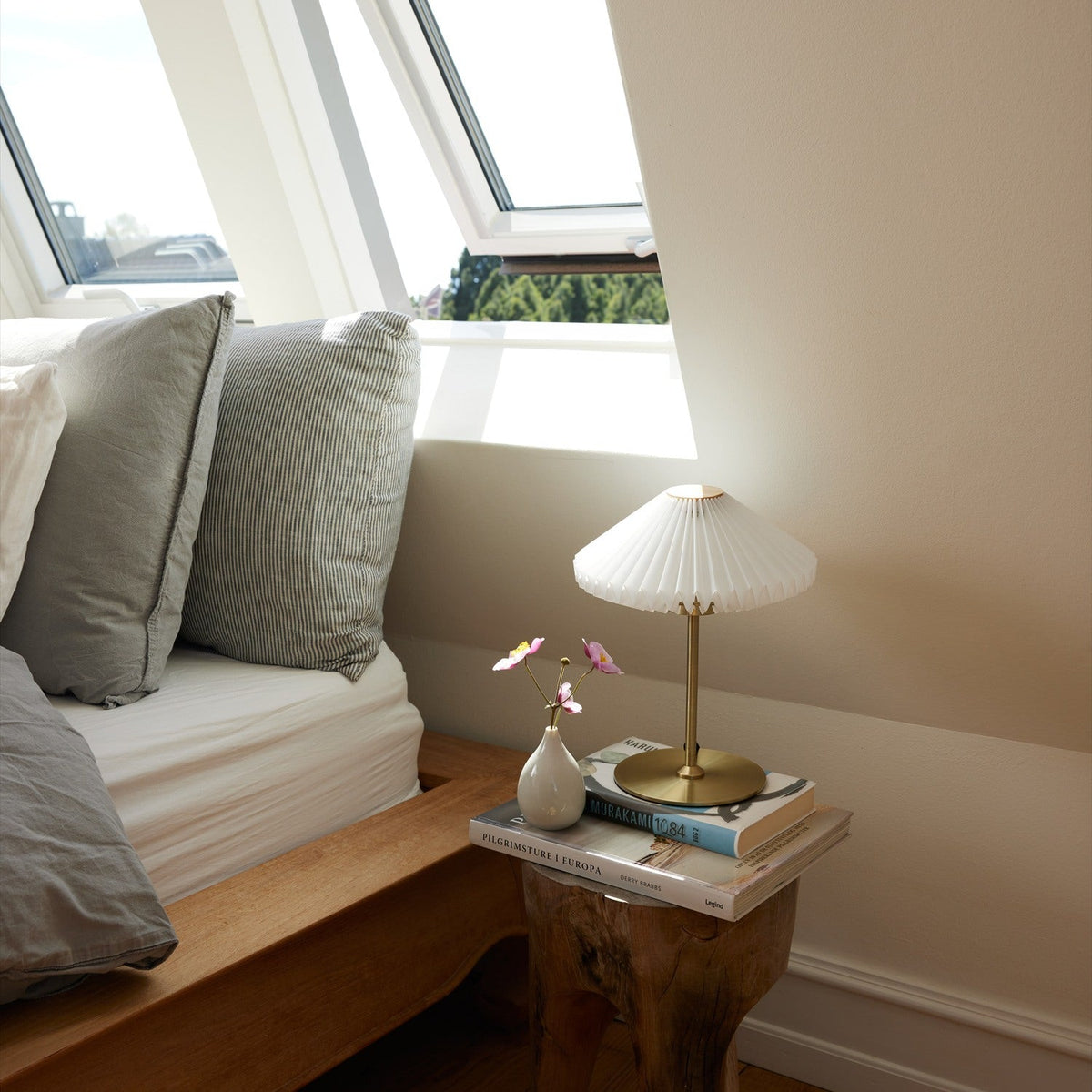 Paris LED Go Rechargeable Table Lamp with pleated white shade and slim antique brass stem, styled on a stack of books beside a bed near a sloped window.
