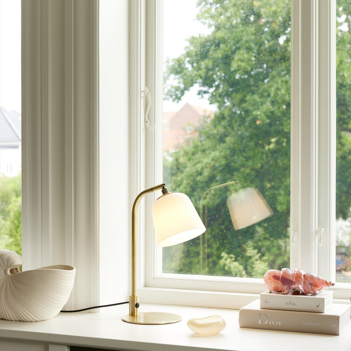 Room table lamp in opal glass and antique brass with a bell-shaped shade on a curved arm and round brass base, shown on a windowsill.
