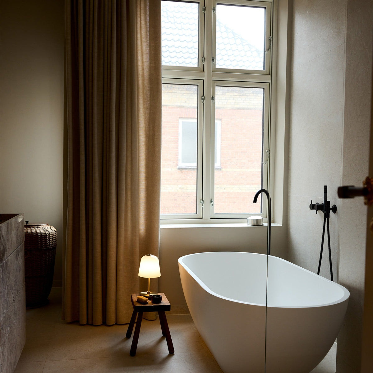 Room LED table lamp with opal glass bell shade and antique brass base, shown lit on a wooden stool beside a freestanding bathtub.