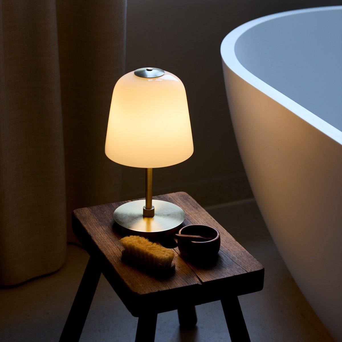 Room LED 2.5W Outdoor Table Lamp in opal and antique brass, bell-shaped glowing glass shade on a slim brass stem with round base, shown lit on a wooden stool beside a bathtub.