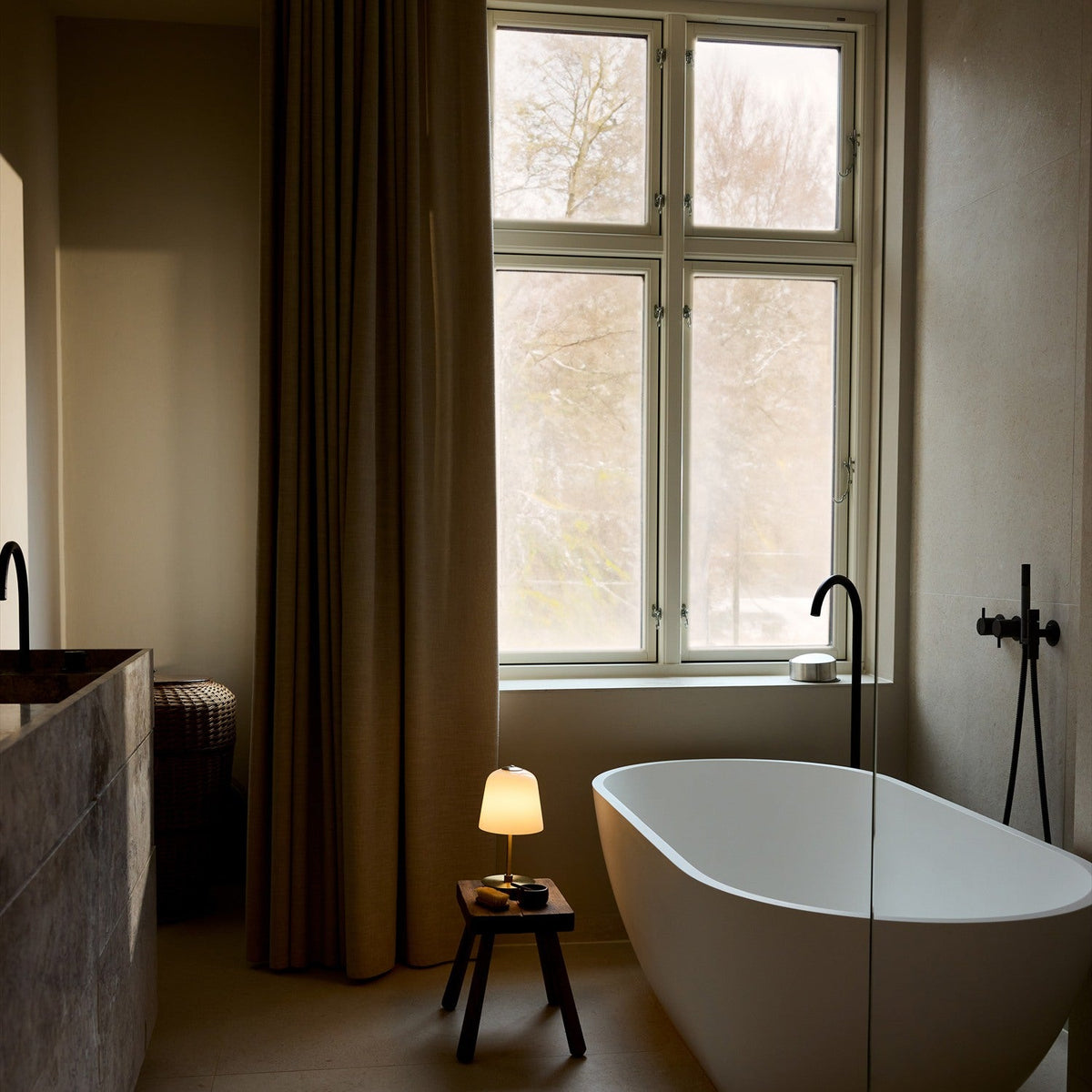 Room LED table lamp with opal glass bell shade and brass stem, glowing softly on a wooden stool beside a freestanding bathtub.