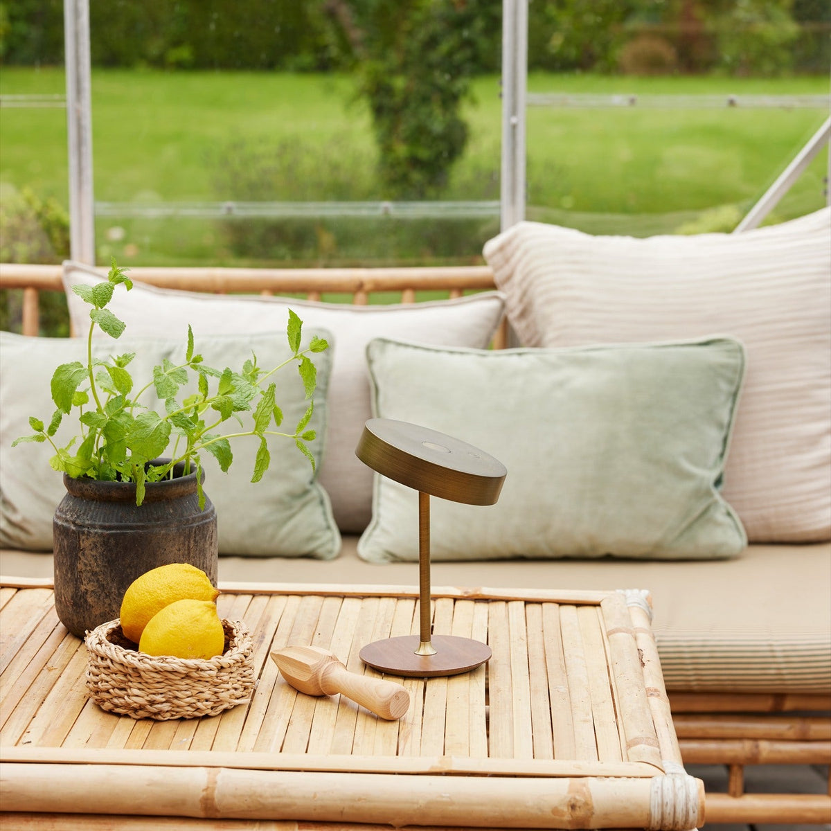 On the Move LED Rechargeable Table Lamp 13cm in antique brass with a round wood base, shown on a bamboo table beside cushions, lemons, and a potted mint plant.