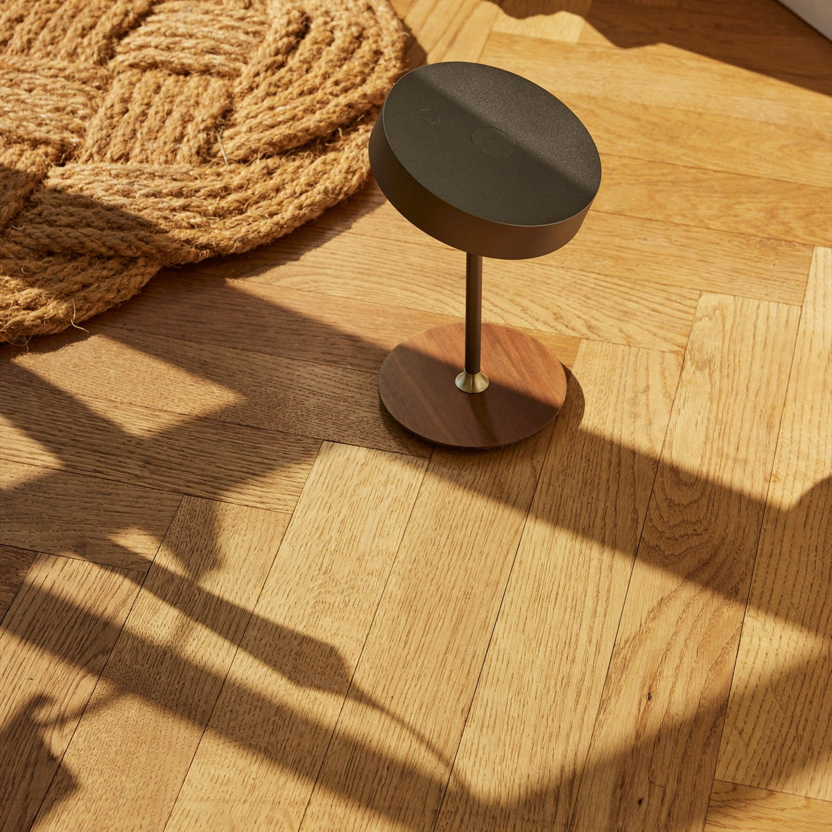 On the Move LED rechargeable table lamp, 13cm, gunmetal drum shade on slim stem with walnut disc base and brass collar, shown on a wooden floor.