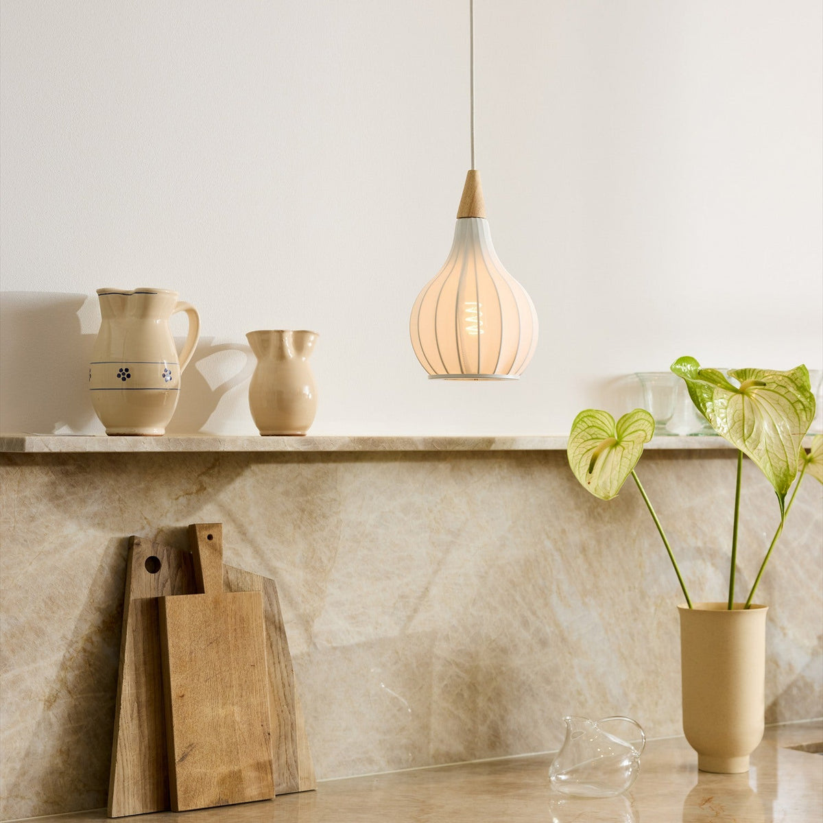 Drops Textile Pendant Light 17cm in white, an onion-shaped ribbed shade with pale wood cap, hanging above a marble kitchen counter with ceramic jugs and plants.