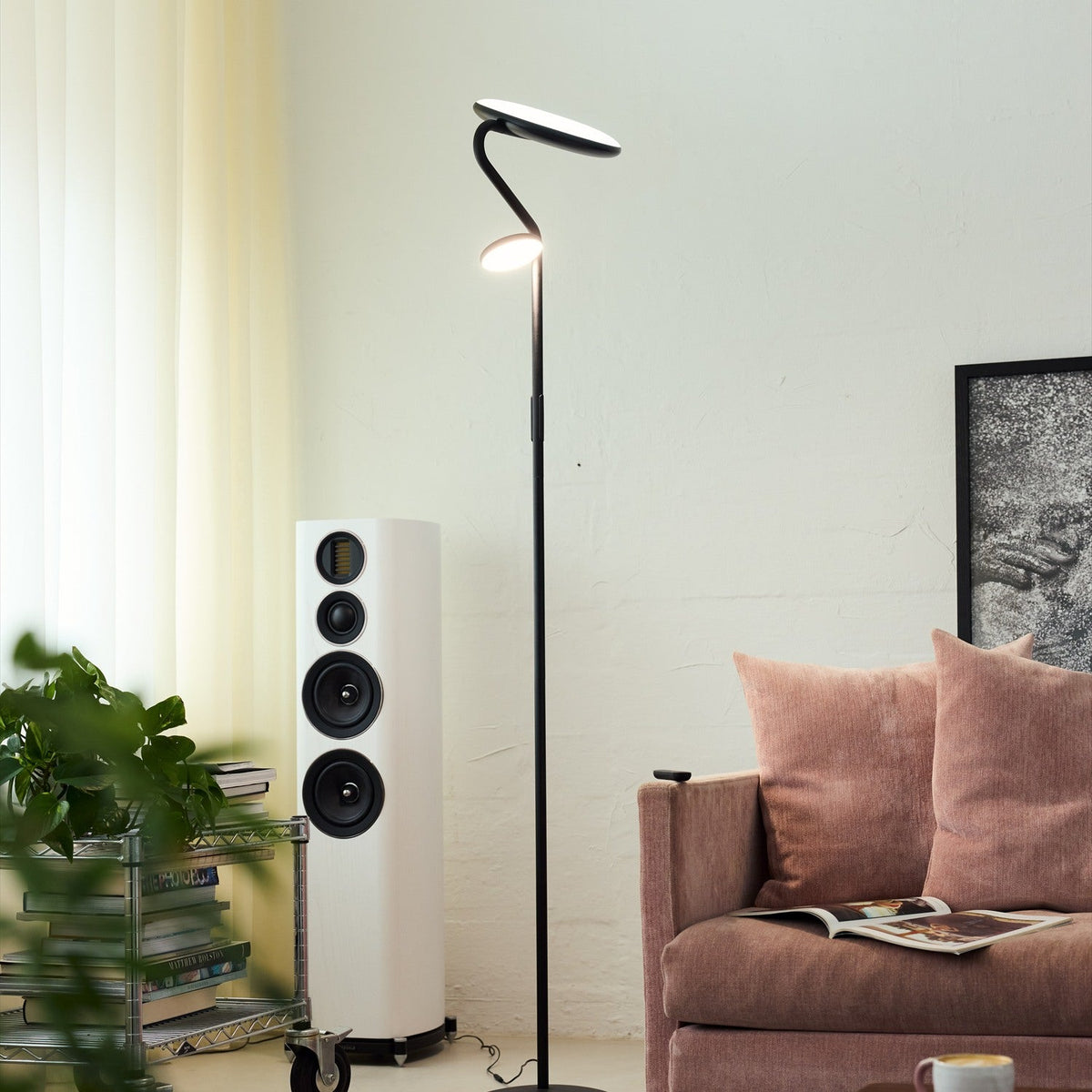 Slim LED Uplight Floor Lamp in black, with a slender curved stem, round base, and two flat disc heads—one upward-facing, one angled down—shown beside a pink sofa in a living room.