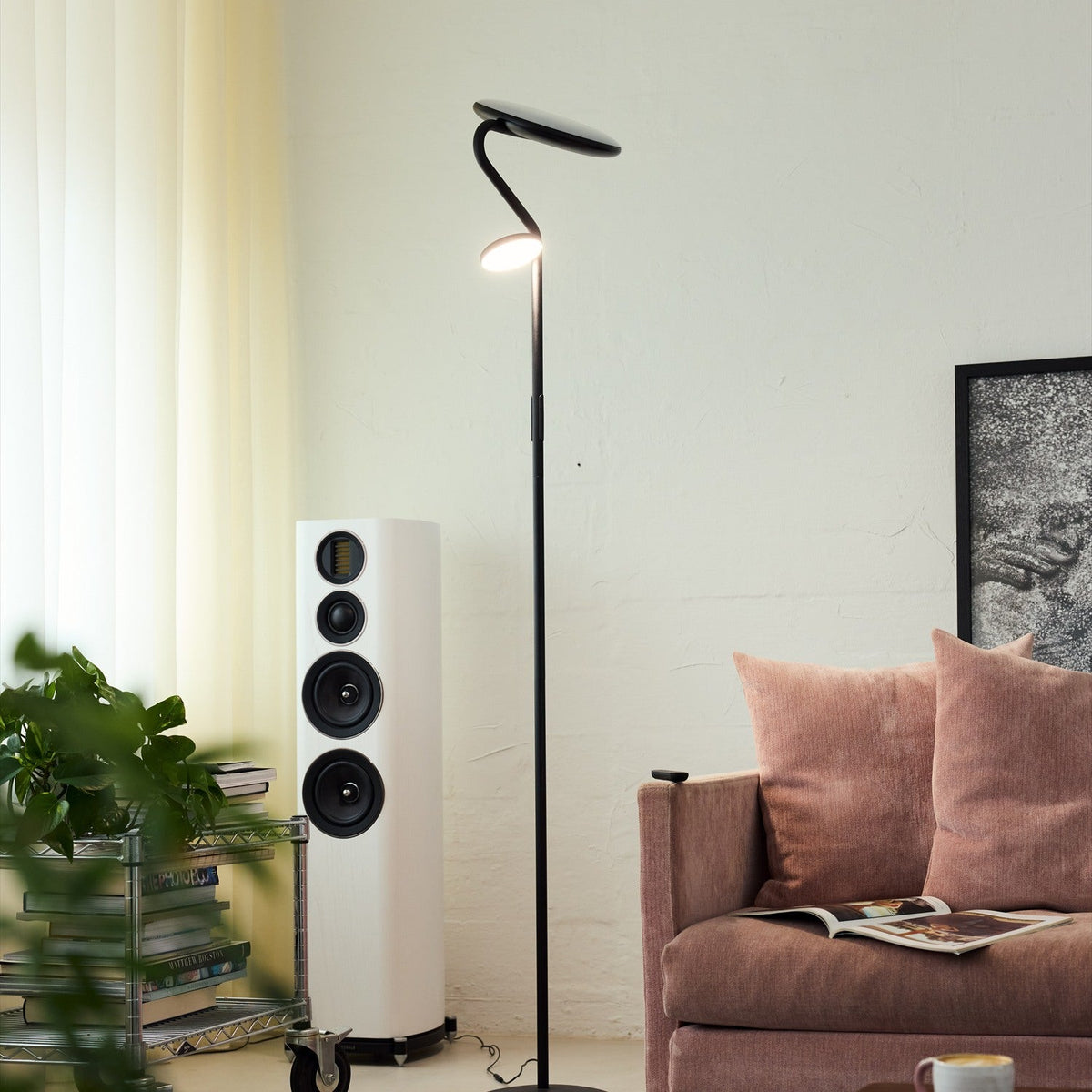 Slim LED Uplight Floor Lamp in black, slim curved stem with round disc base and two flat round heads, shown in a living room beside a pink sofa and speaker.