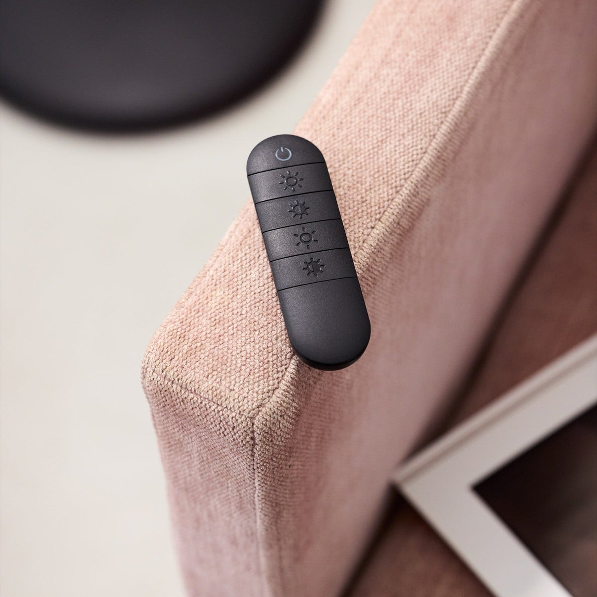 Black remote control with power button and four brightness/dimmer settings resting on a pink upholstered armchair, used to operate the Slim LED Uplight Floor Lamp.