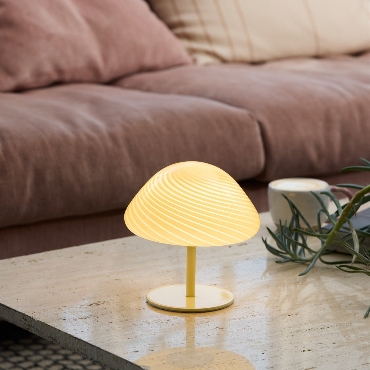 Mini Mush Glass Cable Table Lamp in cream, its ribbed dome shade glowing on a slim black stem, shown lit on a coffee table beside a sofa.
