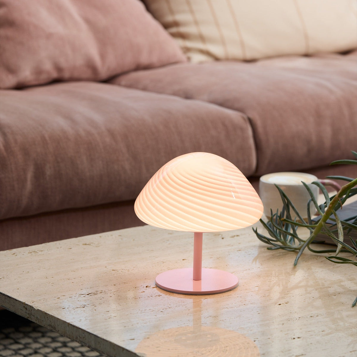 Mini Mush Glass Cable Table Lamp in rose with a swirled, spiral-fluted dome shade on a slim metal stem and round base, glowing on a coffee table beside a pink sofa.