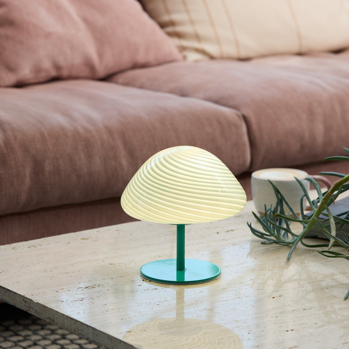 Mini Mush Glass Cable Table Lamp in turquoise with a wide spiral-fluted mushroom dome on a slender green stem and round base, shown lit on a coffee table beside a sofa.