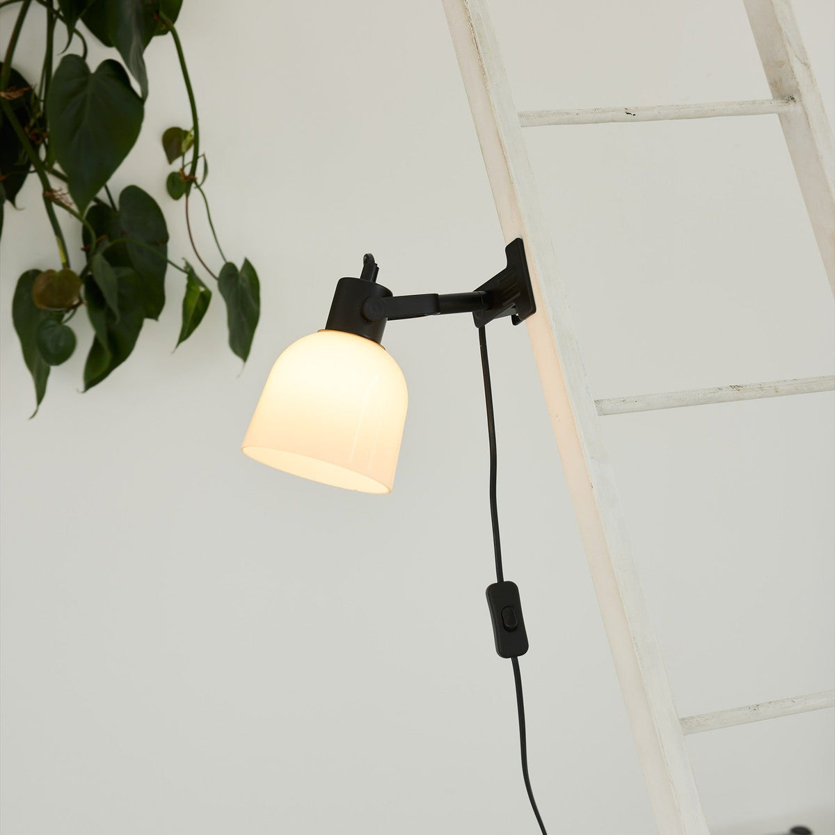 Glow Clip-On Table Lamp in black and white, with an opal glass bell shade on a jointed black arm, clamped to a white ladder rung near hanging plant leaves.