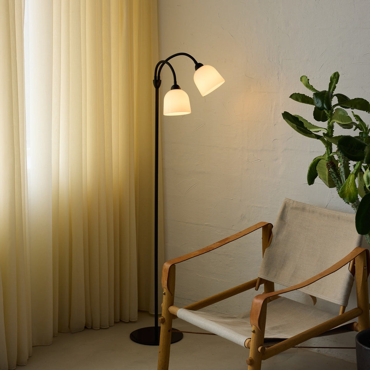 Glow 2 Light Floor Lamp in black and white, with a slim stem and round base supporting two curved goosenecks ending in opal glass bell shades, shown lit beside a chair and plant in a living room.