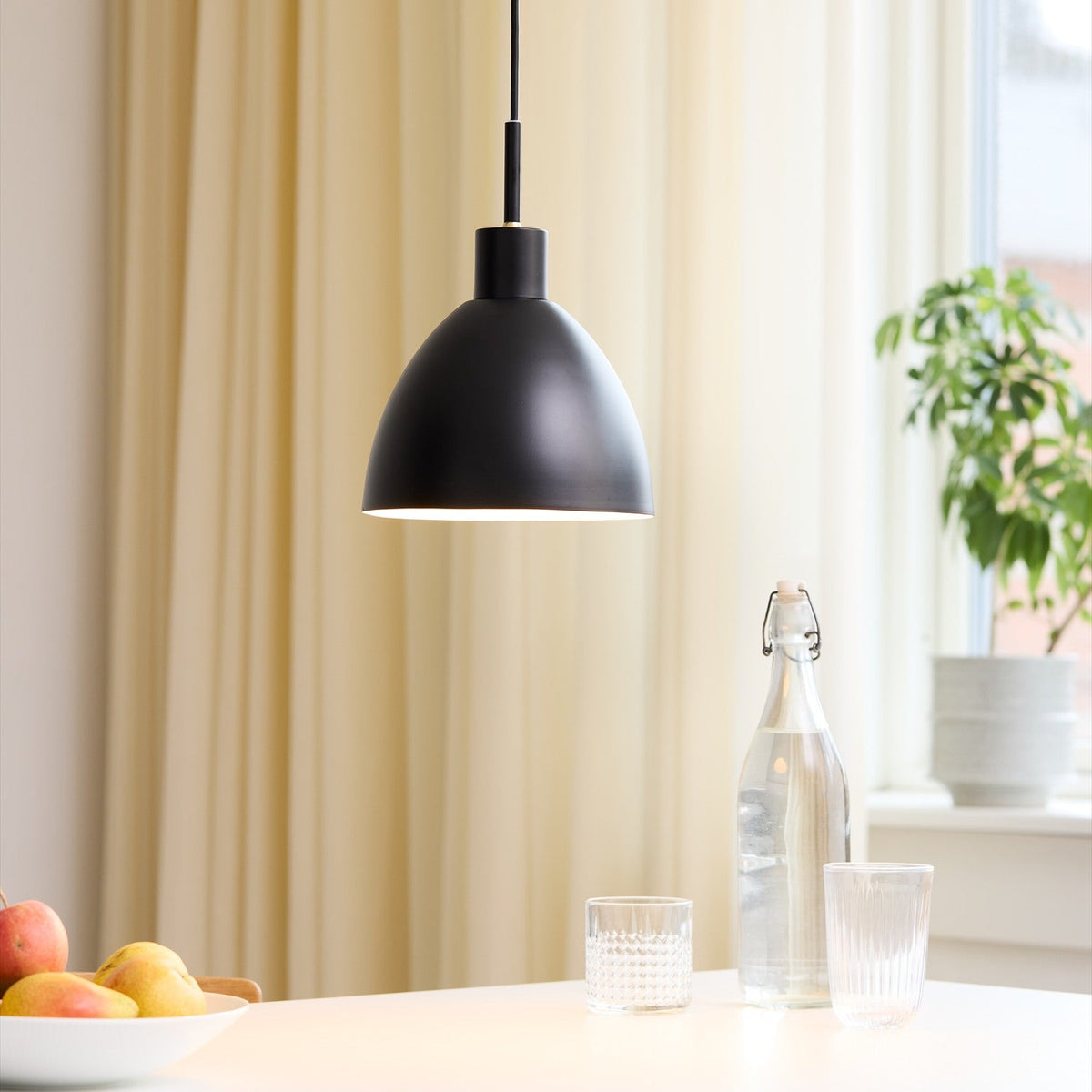 Rock Pendant Light 20cm in black, dome-shaped shade with white interior, hung above a dining table in a room with cream curtains and a potted plant.