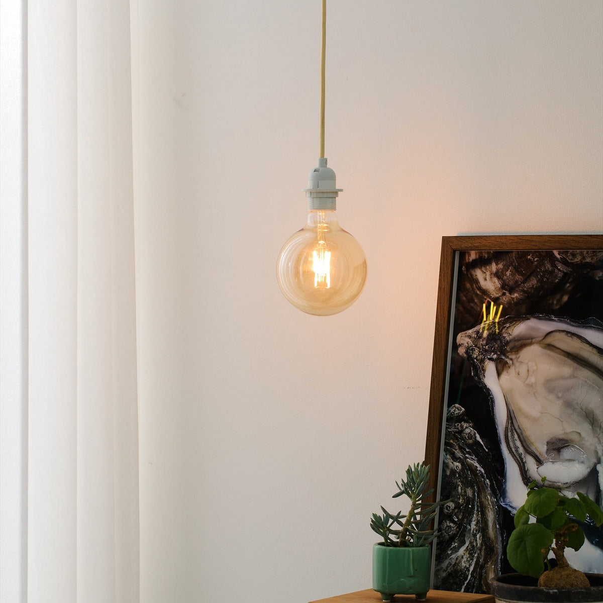 Basic Socket Textile Cable Cord Set 2m in cream, shown as a pendant light with white lampholder and amber globe bulb, hanging near a curtain in a living space.
